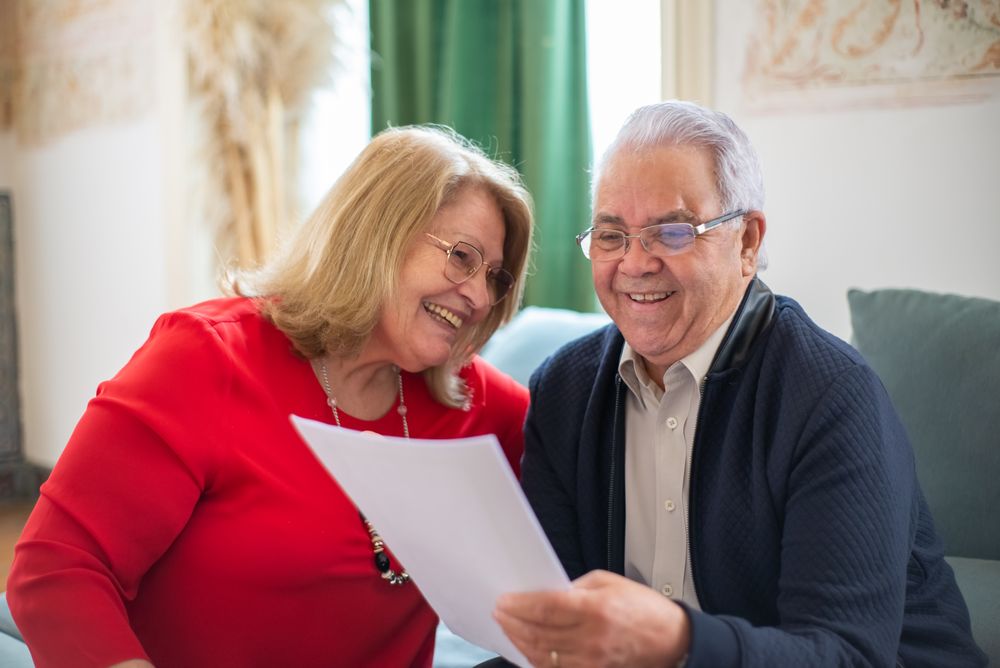 Elderly couple joyfully reading a paper together on the couch in a cozy indoor setting.