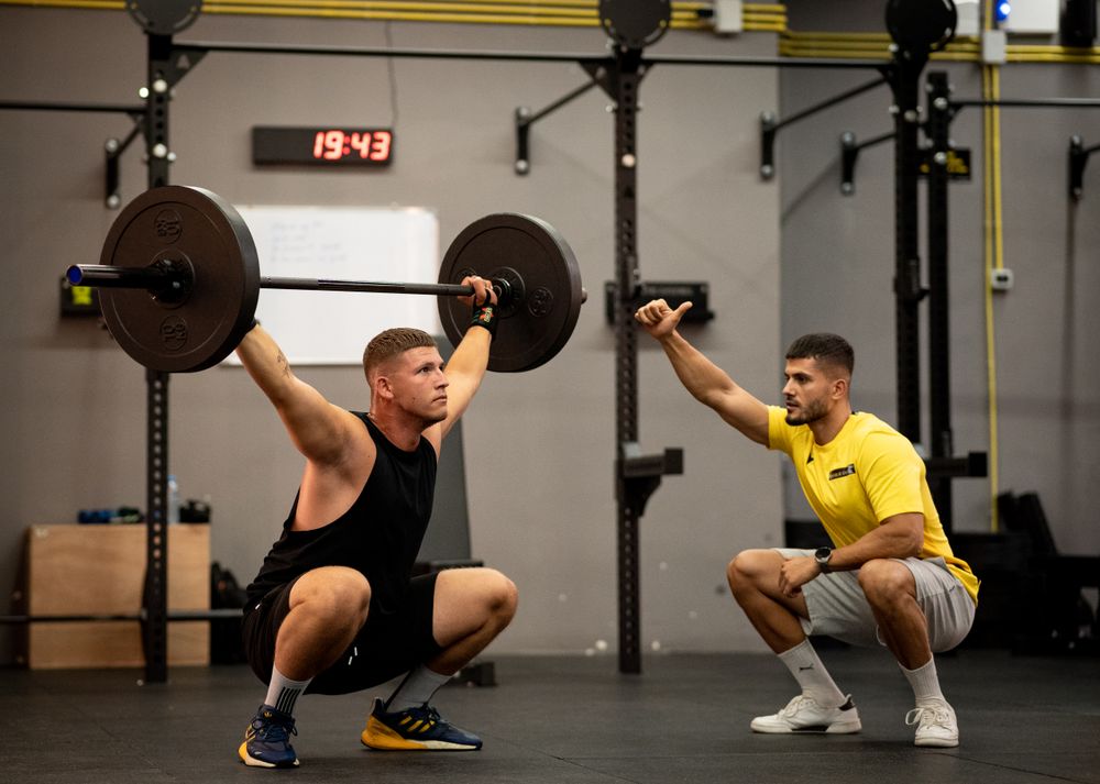 Man lifting weights under coach supervision in a gym setting, focusing on strength training.