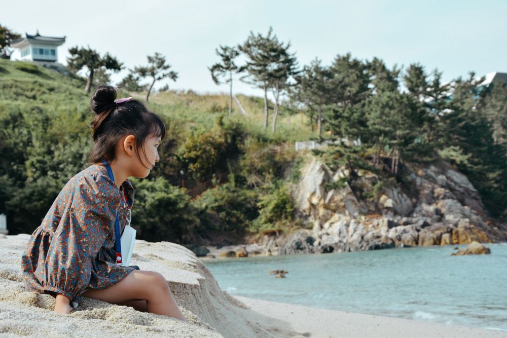 A young girl sitting on a sandy beach in South Korea, gazing at the ocean.