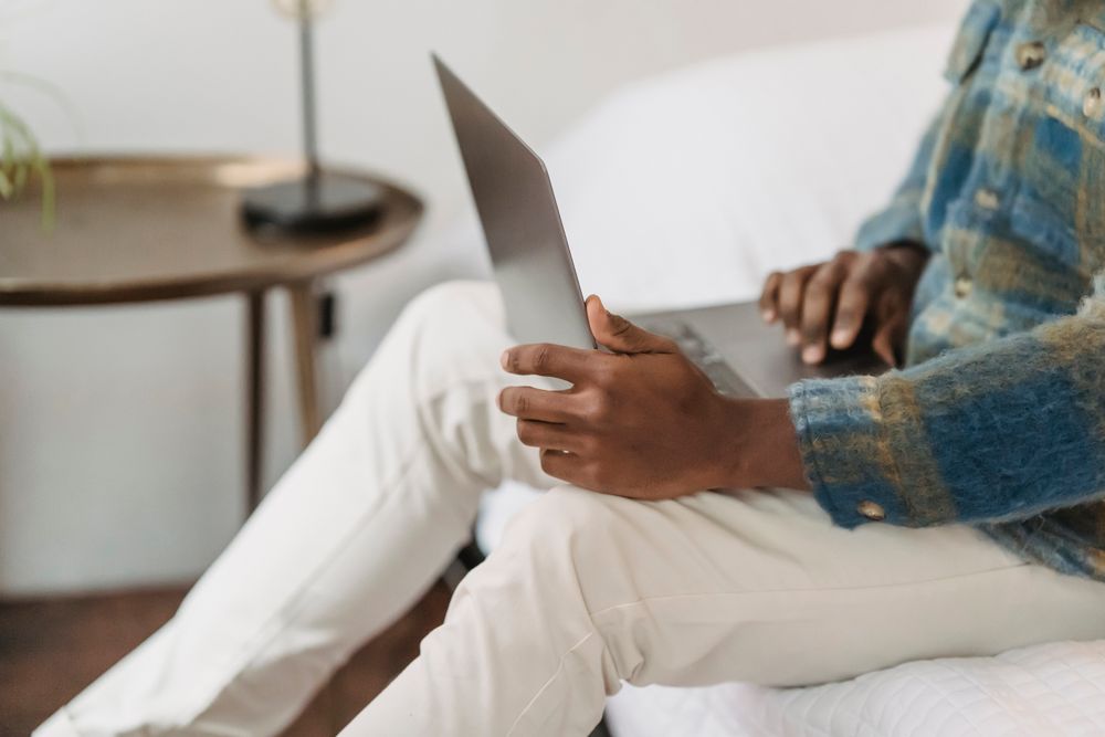 Side view of crop faceless African American male sitting on comfortable sofa and surfing internet on portable computer while remotely working at home