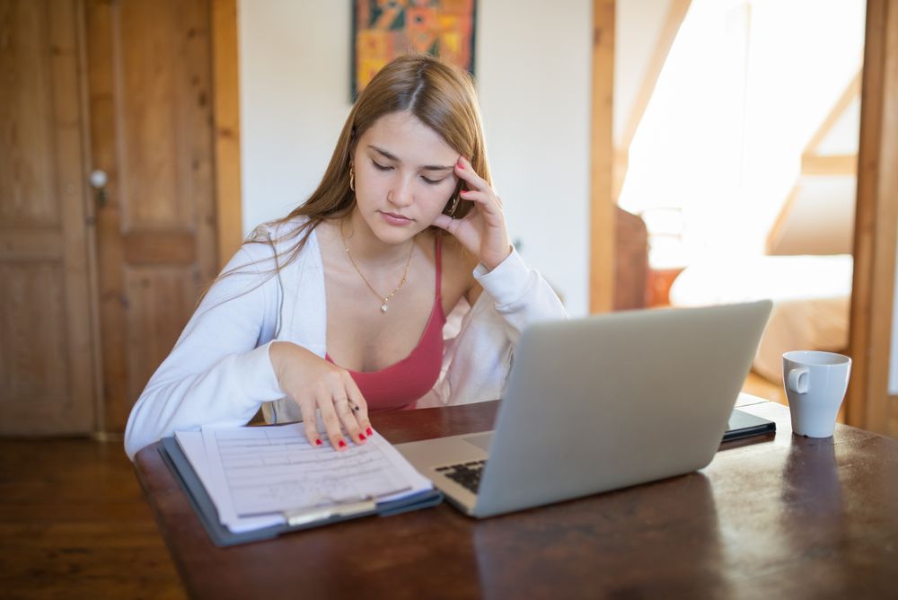 Focused young woman working on documents and laptop in cozy home setting.